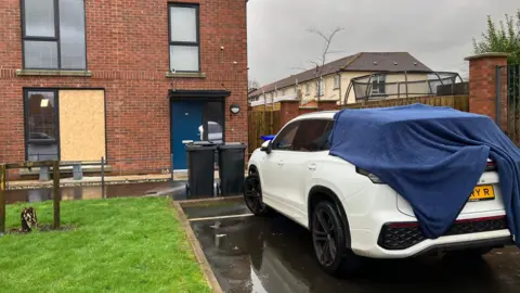 A modern red-brick house with a boarded up window and a white car with a blue blanket thrown over the back of it.