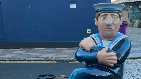 Beryl Cook sculpture of a sailor sat on a bench in the barbican outside of a pub. The sailor is wearing a navy uniform and a white and blue hat with his arms crossed. Blue wall in the background, with pebbled street to the right.