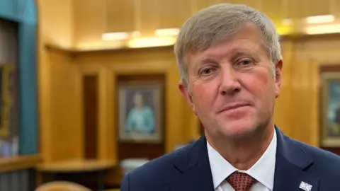 Peter Harris looking at the camera while standing in a wood-panelled council chamber. He is wearing a suit and tie. The tie is maroon and the blazer is navy blue. There are portraits on the wall in the background. He has two metal pin badges on a lapel - one of the union flag and one of the Essex triple seaxes.
