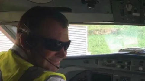 Griffiths sitting in a helicopter wearing a fluorescent jacket, dark glasses and is wearing a headset microphone. 