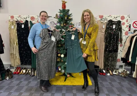 BBC Siobhan Shaw with dark hair on the left wearing blue and Lizzie Dibble with blonde hair is on the right wearing yellow. They are both holding dresses, while other dresses and a Christmas tree are behind them.
