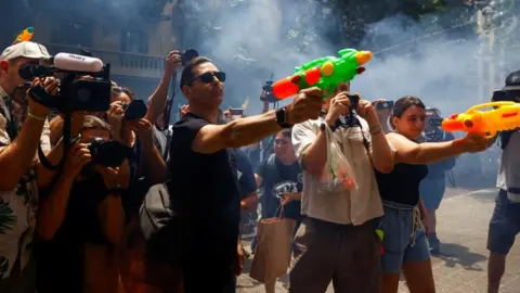 Reuters vīrietis un sieviete, kurai ir ūdens pistoles, savukārt vairāki cilvēki ar video un joprojām kamerām tos filmē protesta laikā. Aiz viņiem gaisā karājas dūmi.