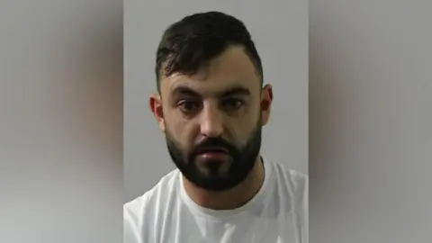 Mugshot of Kristofer Mawhinney. He has short dark black hair and a black beard and is wearing a white t-shirt