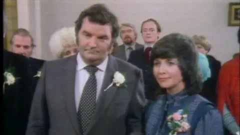 Lisabeth Miles as Megan Harries, wearing a blue dress with a floral cortege, on her wedding day in 1988, she is stood next to a man in a dark grey suit. There are people behind them. It is their wedding day with guests stood with them.