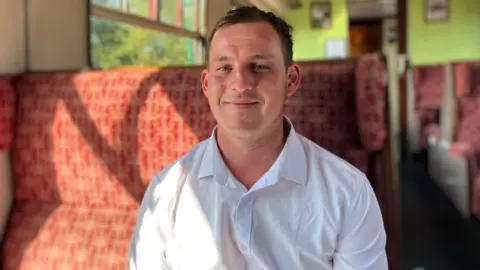 Henry Godfrey-Evans/BBC Robert Bentley has short dark hair and is wearing a white shirt. He is smiling and sitting in the exact same old-fashioned seat as Sandra Bullock and Nicole Kidman.