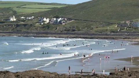 BBC Weather Watcher/Poppy The view across Croyde Bay. The water is busy with swimmers and surfers while the beach is busy with walkers. There is an offshore wind creating surf. There are houses on the opposite headland.