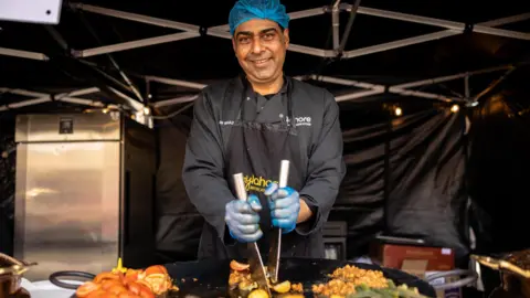 Bradford Council A man in black chef's clothing stands in a gazebo and is cooking on a huge pan