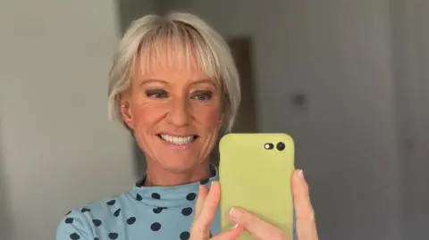 Ruth Dodsworth A woman with short light blonde hair and several silver ear piercings. It is a saelfie image image in front of a mirror. She is wearing a blue top with black polka dots, is holding a phone in a green case and is smiling.