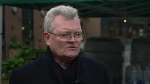 BBC / Peter Whittlesea A man with grey hair and glasses. He is wearing a black coat and standing in front of a building.