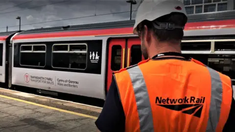 The back of a man with an orange reflective jacket and white helmet looking at a white, red and black train at a platform