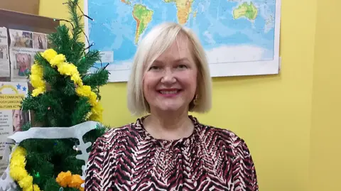 Mary Lafferty with short blonde hair and is wearing a red, black and white patterned top. Behind her is a Christmas tree and world map on a yellow wall.