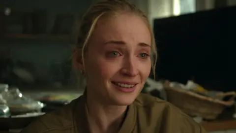 Picture of Sophie Turner looking upset, with tears in her eyes and her hair pulled back. She is wearing a khaki bomber jacket that is slightly unzipped. Messy blurred household items appear in the background.