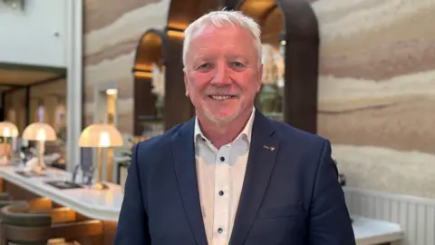 Charlie Le Noury/BBC Lee Madden has white hair and stubble on his chin and is wearing a navy jacket and white shirt with black buttons.
he is standing in front of a long table with lamps on it.
