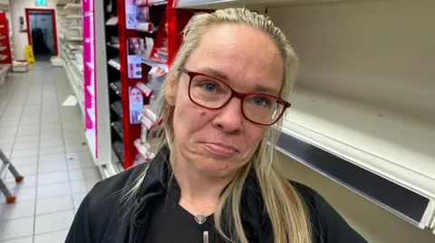 Leighann Holden - a woman with long, blonde hair looks sad at the camera. She is posing in front of empty shelves and is wearing a pair of red, square frame glasses, a black jacket and a black t-shirt.