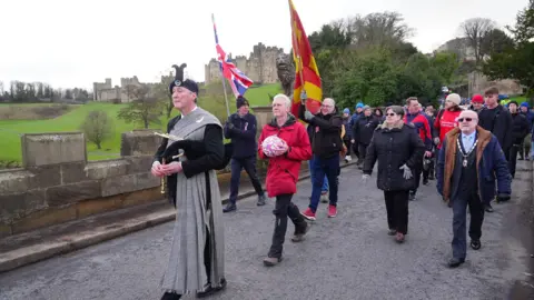 PA Media A Northumberland piper leads a procession. A man in a red coat carries the ball. A mayor is also there and wears a gold chain around his neck. Alnwick Castle is in the background.