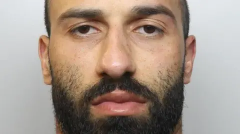 A mugshot of a young man with short hair and a dark beard. He is staring directly at the camera.