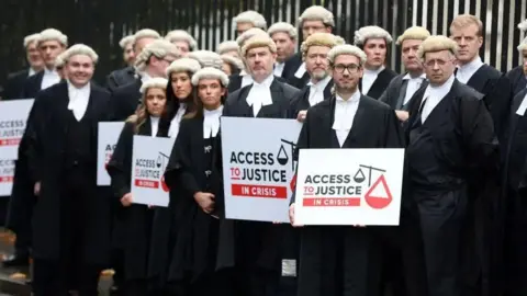 Pacemaker A number of barristers, both male and female, standing in black robes and white wigs, a number of them holding placards saying access top justice in crisis