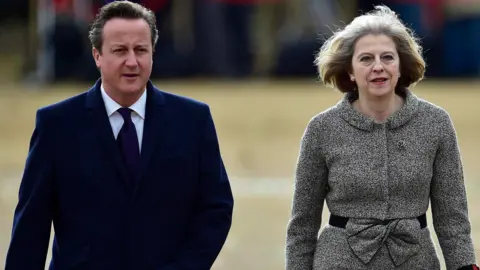 PA Media Lord David Cameron in a blue overcoat, white shirt and blue tie, walks alongside Baroness Theresa May, who wears a grey and white patterned jacket with a large bow belt