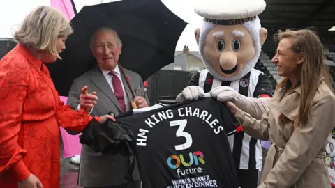 PA Media King Charles is presented with a Grimsby Town shirt with the lettering "HM King Charles 3 Our Future Winner Winner Chicken Dinner". The shirt is being presented by the club mascot The Mighty Mariner. There are two women also involved in the presentation. 
