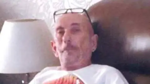 family handout Man wearing a white t-shirt, sitting a in brown leather chair next to a white lamp, with glasses on his head