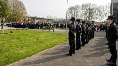 Kent Police Uniformed police officers stand round a green square, some with their heads lowered as they pay respects.