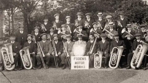 Foden's Band An old black and white photograph of a brass band. Members are wearing hats, suits and holding instruments. One of them is holding a large shield award and is sitting behind a sign that says Foden's Motor Works.