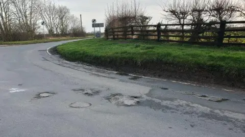 A well-worn section of rural road between Brandesburton and North Frodingham, showing evidence of potholes and patching work. Next to the road is a grass verge and hedgerow.