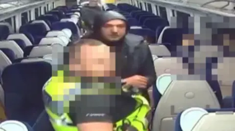 Prata is wearing a black jacket with the hood up as he is arrested on a train by a uniformed police officer whose face we have blurred out