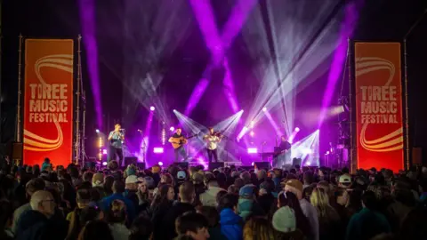 Tiree Music Festival A large audience gathers in front of a brightly lit stage at the Tiree Music Festival, with performers playing under dramatic purple and white spotlights.