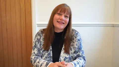 Supplied Sharon Nelson is sitting at a desk and smiling at the camera. She has long brown hair and is wearing a blue and white jacket over a black top.