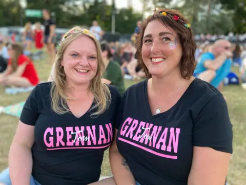 Alex Pope/BBC Two women stand next to each other in a field full of people behind them.
They wear dark-coloured T-shirts with the words Tom Grennan in pink and white. Both have flowers in their hair and are smiling. 