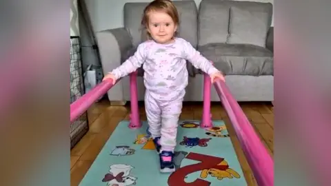 A young girl wearing pink patterned clothes is in a living room. She has her hands on two parallel pink bars which are attached to a walking mat. In the background is a grey sofa. 