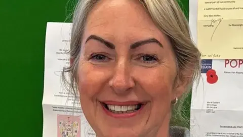 Vicky Pudney A smiling Vicky Pudney looking straight at the camera. She is standing in front of a green noticeboard with several letters pinned to it.