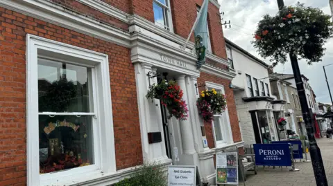 Elliot Deady/BBC The front of Witham Town Hall, where there are hanging baskets with flowers at the front door and a flag above the entrance.