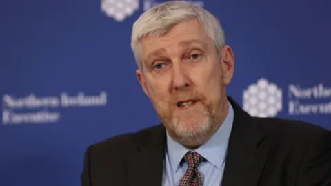 John O'Dowd has short grey hair and a grey and ginger beard. He is wearing a dark suit, blue shirt and red and green patterned tie. He is standing in front of a blue blurred background.