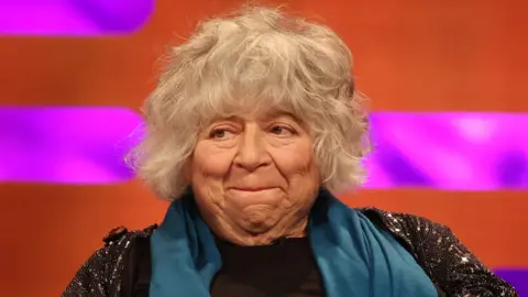 Miriam Margolyes - a woman with medium-length grey hair, a blue scarf and a dark top - looks to her right and smiles as she appears on the Graham Norton Show.