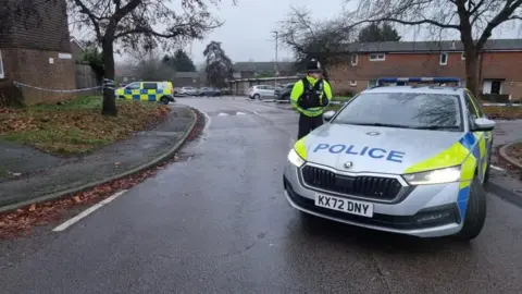 Dawid Wojtowicz/ BBC A residential street with two-storey brick houses on either side. There are paths and trees at the side of the road. There is a grey police car in the foreground and an officer in black helmet and yellow jacket is standing beside it. A police van is visible in the distance.