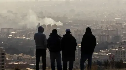 Four men stood on a hill looking at smoke over Tehran
