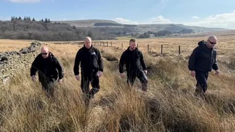 Four men dressed all in black walk through long, dry grass in between a wire fence and stone wall through a sloped field. Moorland and forest can be seen in the distance on a bright, sunny day. 