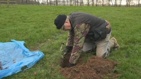 David Stuckey David Stuckey is using his gloved hands to retrieve fragments from a hole in the ground. He is kneeling in a field and is wearing a woolly hat, spectacles, a camouflage jacket and protective knee pads. There is a piece of blue plastic near the hole on to which he is placing fragments from the hole.