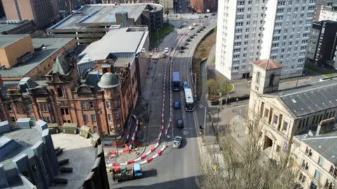 A drone camera view of Cowcaddens Road. Part of the street is taped off to allow road repairs and work to take place. Various cars and buses are going along other lanes. A large red brick building is next to the road, while a white coloured block of flats is also visible.