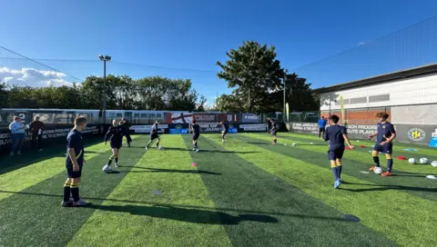 BBC/Harry Low Children on green astro turf pass footballs
