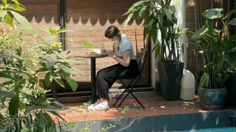 Ethereal Jewellery Leah sits at a table by a swimming pool, writing in a notepad. She has on black trousers, a green top and sandals. There are bushes and plants around her.
