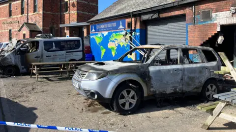 The remains of a Collingwood Seafood-branded van stands outside the shop following a fire. A similarly destroyed car has been abandoned outside the shop. with its rear facing the building. The wall behind it has been knocked down. The building is a single-storey brick construction with a large, central metal garage-style shutter and a pitched, tiled roof. There is a mural to the left of the shutter depicting a map of the world, below a blue sign showing the company name. A line of blue and white police tape is in the foreground.