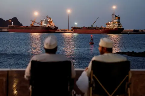 Reuters The Galaxy Globe bulk carrier and the Luojiashan tanker sit anchored on 9 March 2026. In the foreground, in soft focus, two men in white robes sit in chairs overlooking the waterway and the ships. It is dusk and lights from the ship light up the sky. Photo by Benoit Tessier
