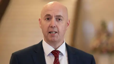 PA Media Jon Burrows is wearing a navy suit, white shirt and red tie. He has no hair and the background is blurry. 