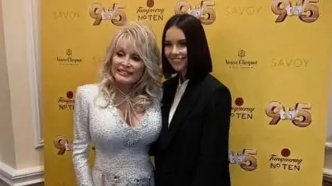 Dolly Parton has curled blonde hair and is wearing a white, silvery dress. Carlin has a straight black bob and is wearing a black suit. 