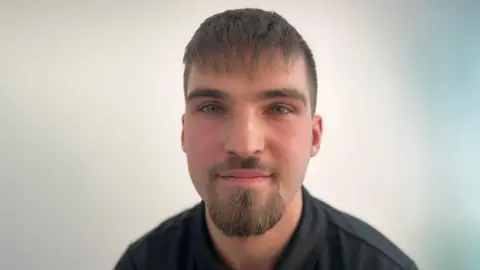 Emma Baugh/BBC Szymon Olszewski is wearing a dark blue polo shirt and is smiling at the camera, behind him is a plain white wall