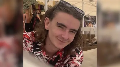 Jamie Collins wearing a black, red and white patterned shirt and sunglasses on his head. He has shoulder length brown hair and piercings in his ears, eyebrow and nose. He appears to be sitting underneath a white marquee on a sunny day.