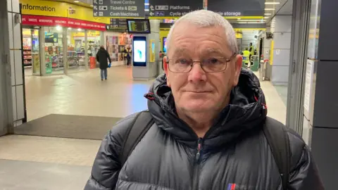 Steve Carey has short grey hair and is wearing dark rimmed glasses. He is standing inside Liverpool Central station and is wearing a grey winter coat and has a rucksack on his shoulders.